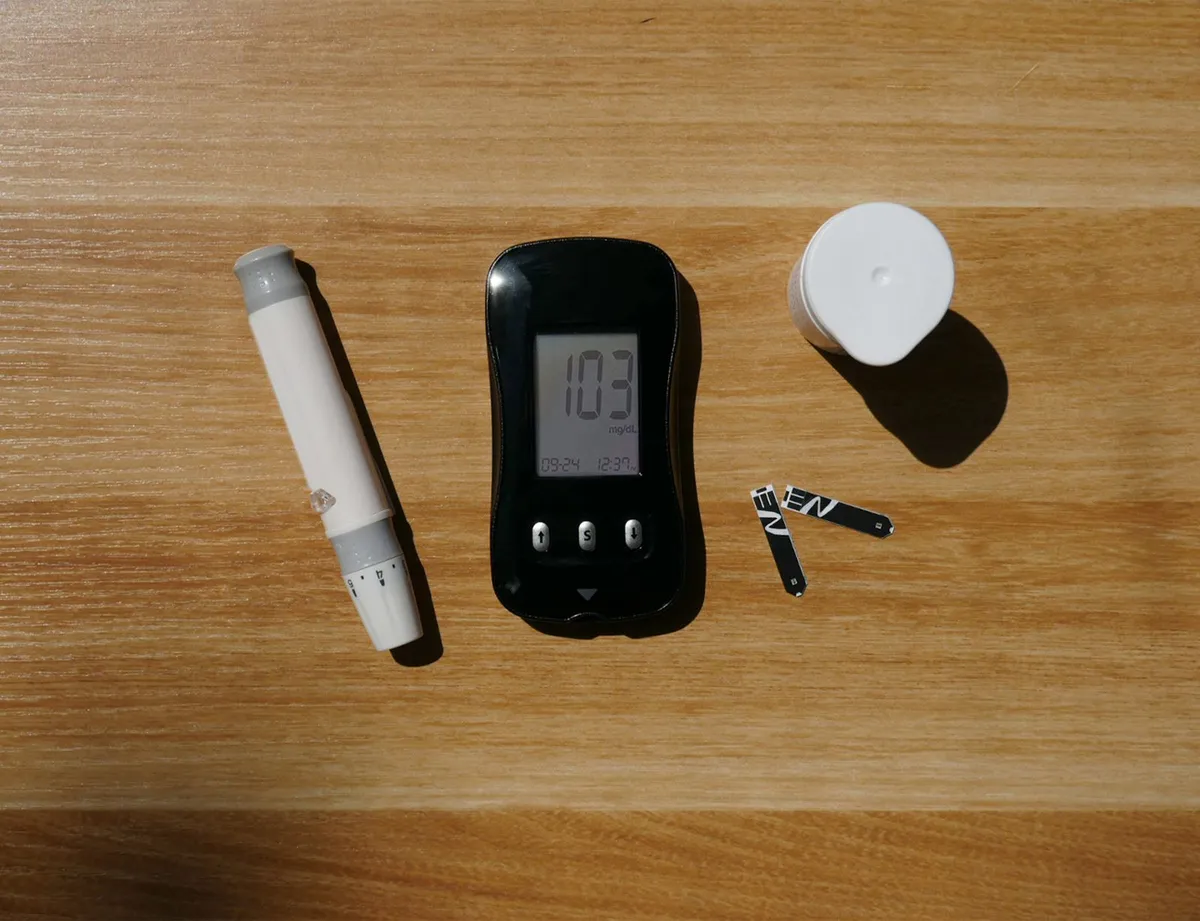 Glicosímetro marcando 103 ao lado de lancetador e tiras sobre mesa de madeira, glicemia na faixa de pré-diabetes